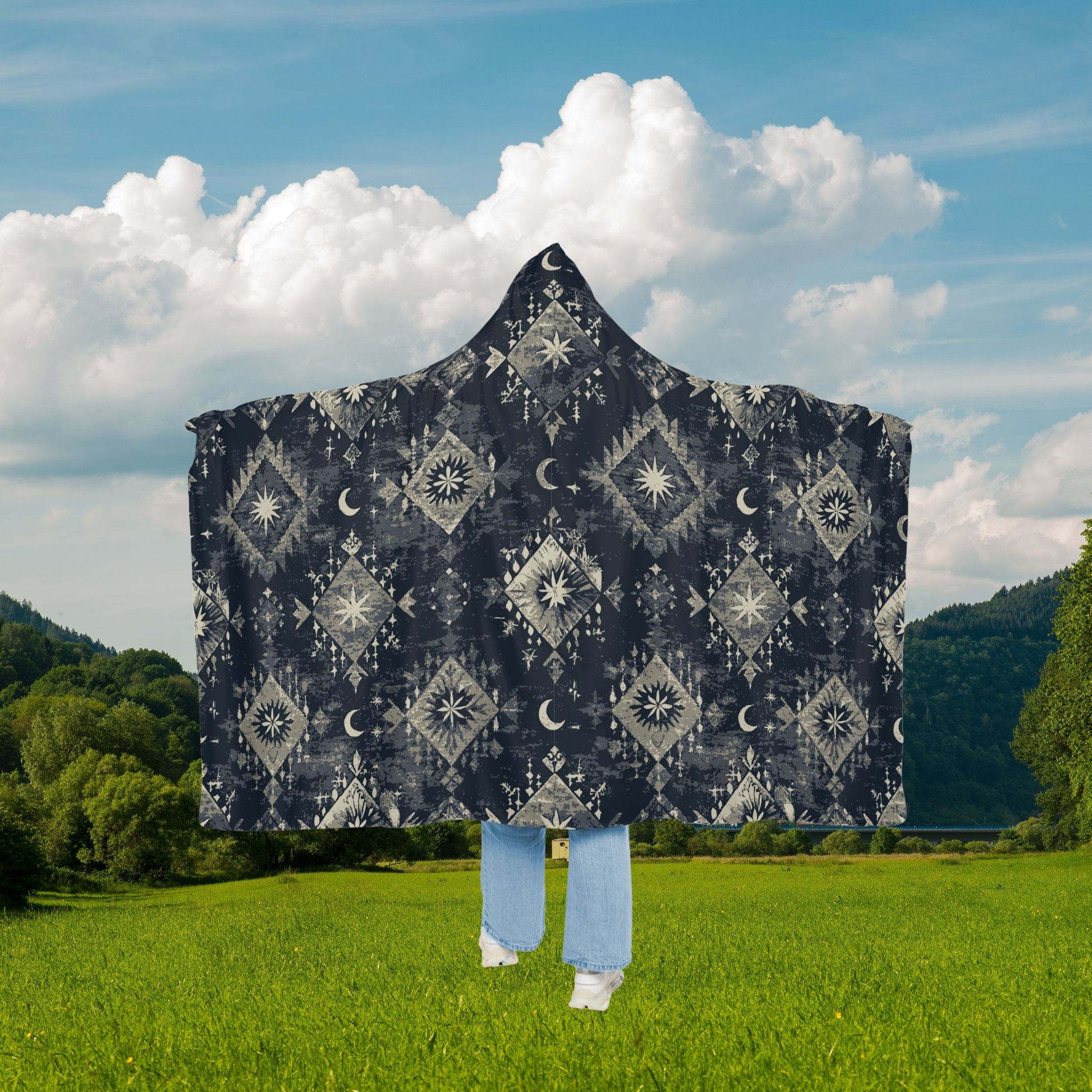 Person wearing a patterned hooded blanket in a green field with a blue sky.