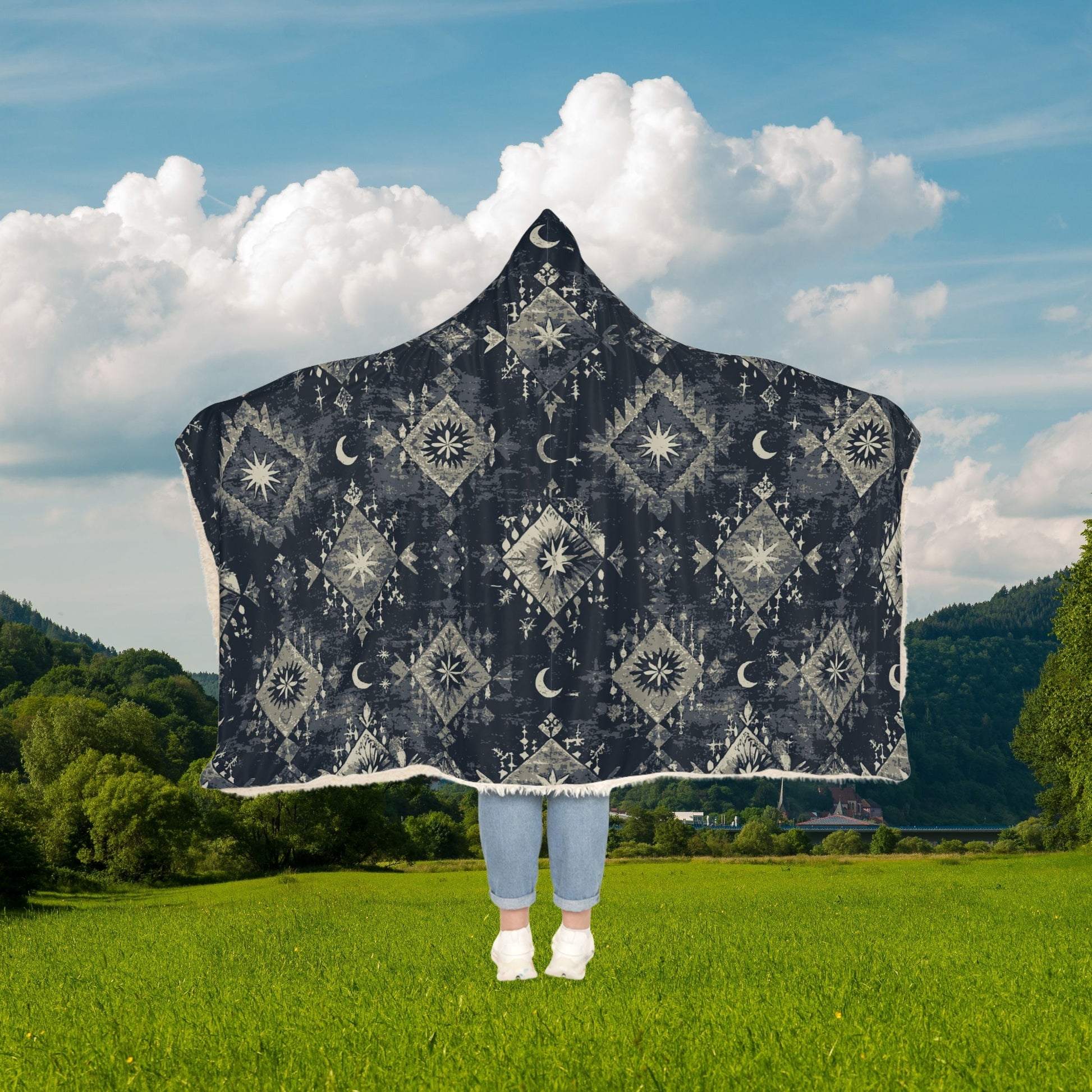 Person wearing a dark hooded blanket with geometric patterns in a green field with trees and blue sky.