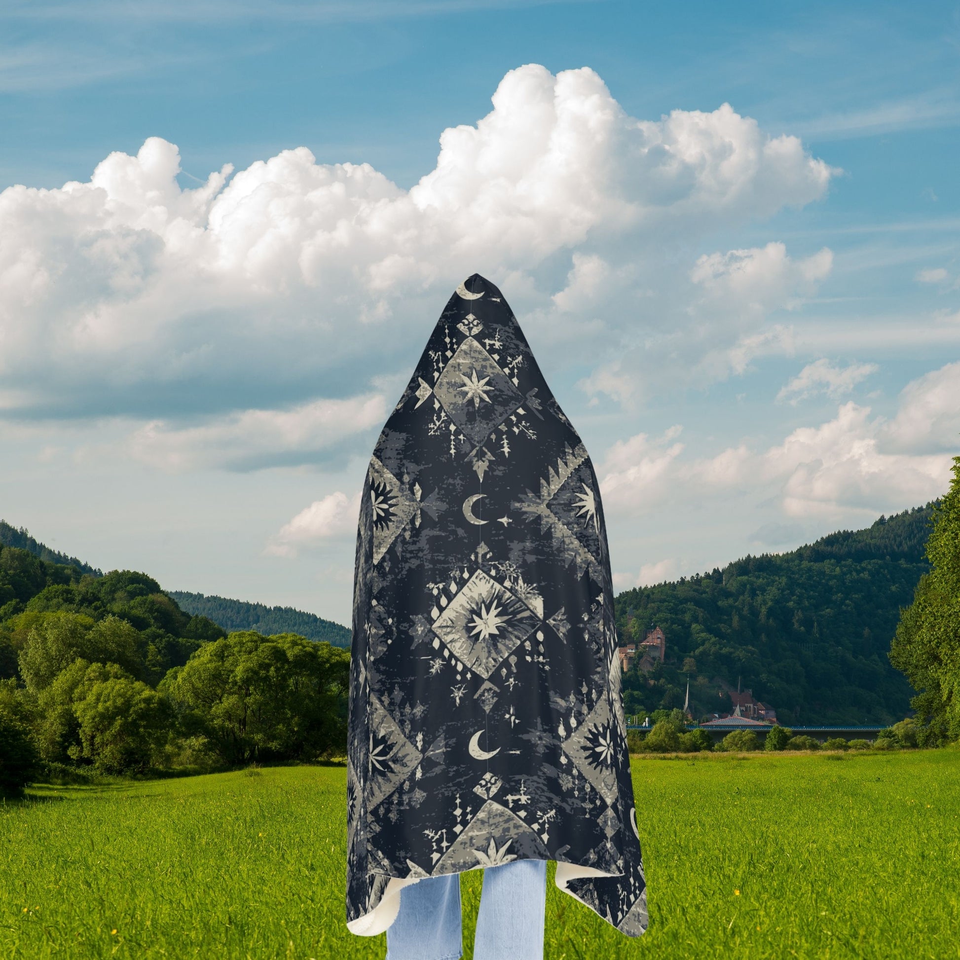 Person wearing a black hooded garment with white patterns in a green field with mountains in the background